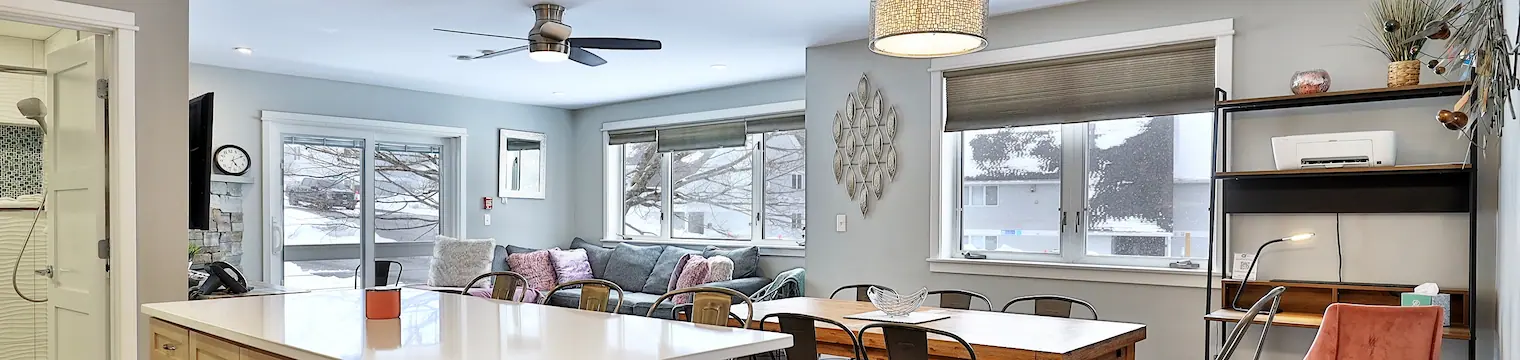 Living & dining room of a nicely-decorated vacation condo with large windows showing a snowy exterior with trees.