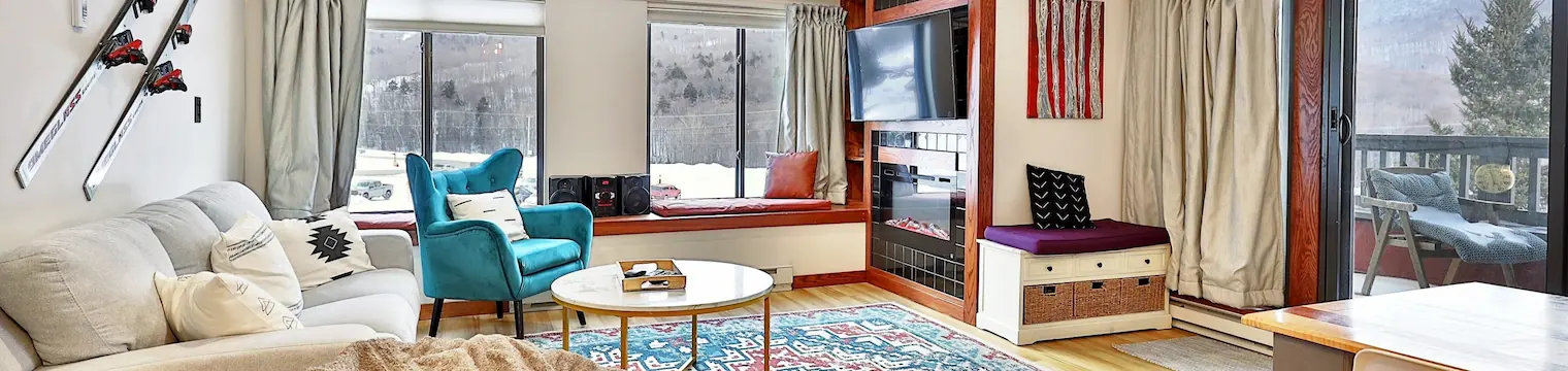 Living room of a vacation property. There's an electric fireplace, a TV, a stylish armchair, skis displayed on the wall, and large windows with a view to snowy woods.