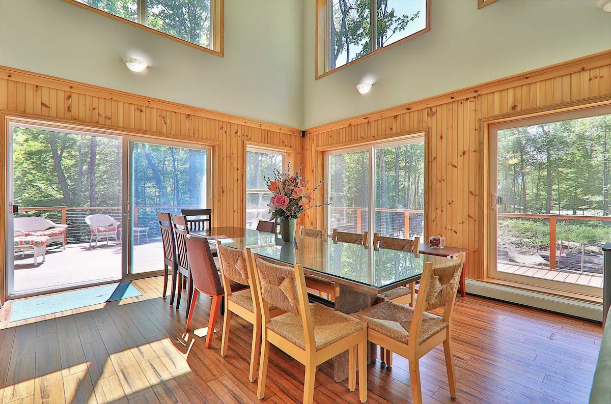 Dining area of a seasonal rental house in Killington, VT. Large windows show a sunny day outside, with plenty of trees and green, seemingly the summer or spring season. 