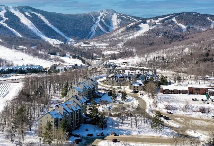 Beautiful drone winter view of Mountain Green Resort condo complex, with its several buildings, cars parked at their fronts, and Killington Mountain in the back.